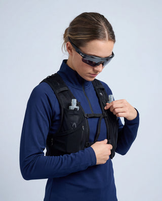Haut technique demi-zip Pavenrod bleu marine Made in France porté par une femme avec gilet d’hydratation de trail et lunettes, fond clair.