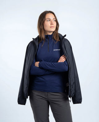 Haut technique demi-zip Pavenrod bleu marine Made in France porté par une femme avec veste coupe-vent sur les épaules, pantalon de rando, fond blanc.