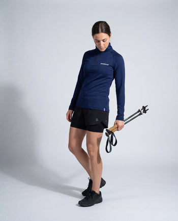 Haut technique demi-zip Pavenrod bleu marine Made in France porté par une femme avec bâtons de randonnée, short et chaussures de trail, fond studio.