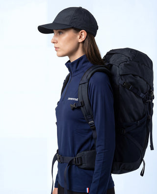 Haut technique demi-zip Pavenrod bleu marine Made in France, vue de profil, porté par une femme avec casquette et sac à dos de randonnée, fond blanc.
