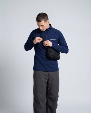 Haut technique demi-zip Pavenrod bleu marine Made in France porté par un homme avec sacoche bandoulière, pantalon outdoor gris, fond studio.