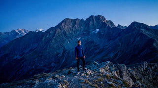 Homme au sommet portant un haut technique zippé Pavenrod bleu nuit, panorama de massif montagneux à l’aube.