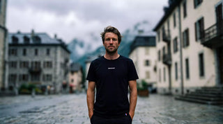Homme portant le t-shirt noir Pavenrod en coton bio, fabriqué en France, en village alpin avec les montagnes en arrière-plan.