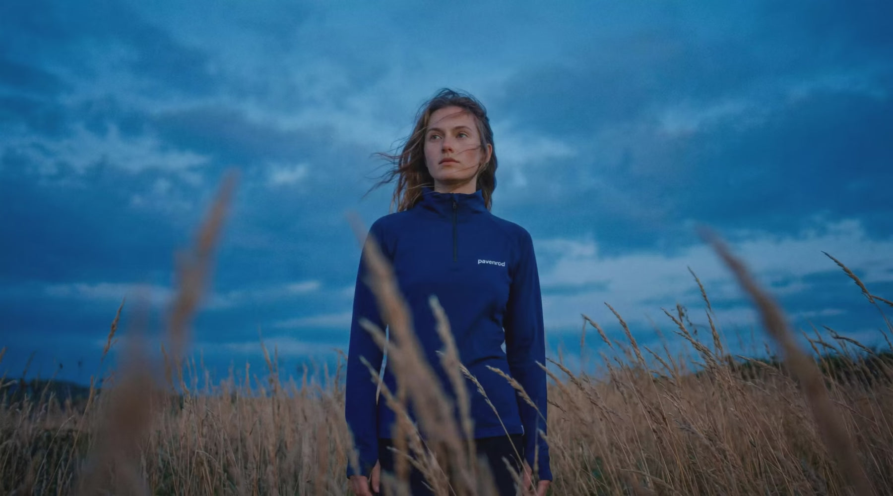 Femme portant un haut technique manches longues zippé Pavenrod, dans une prairie sous ciel chargé à l’heure bleue.